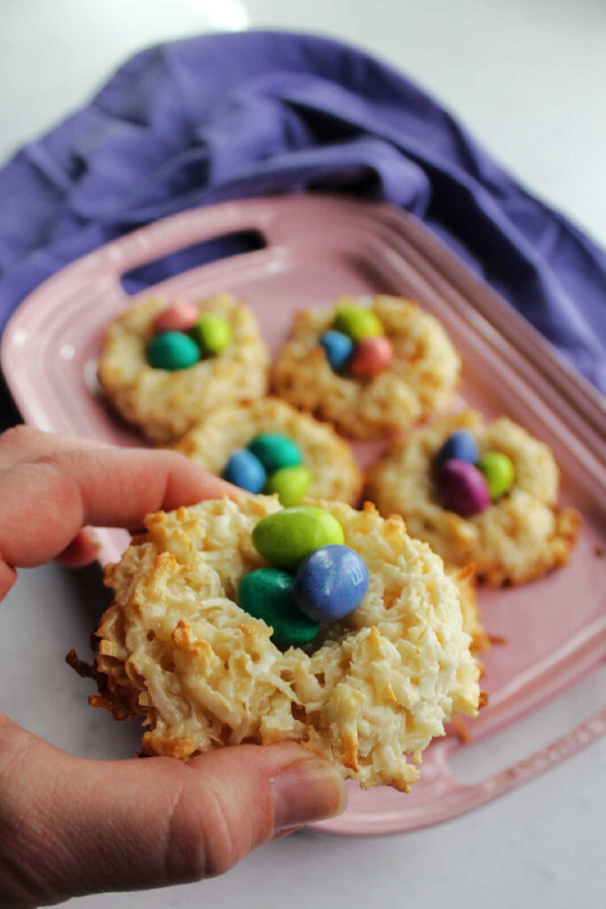 Hand holding coconut macaroon nest with bits of golden brown coconut on the nest and chocolate eggs inside. 