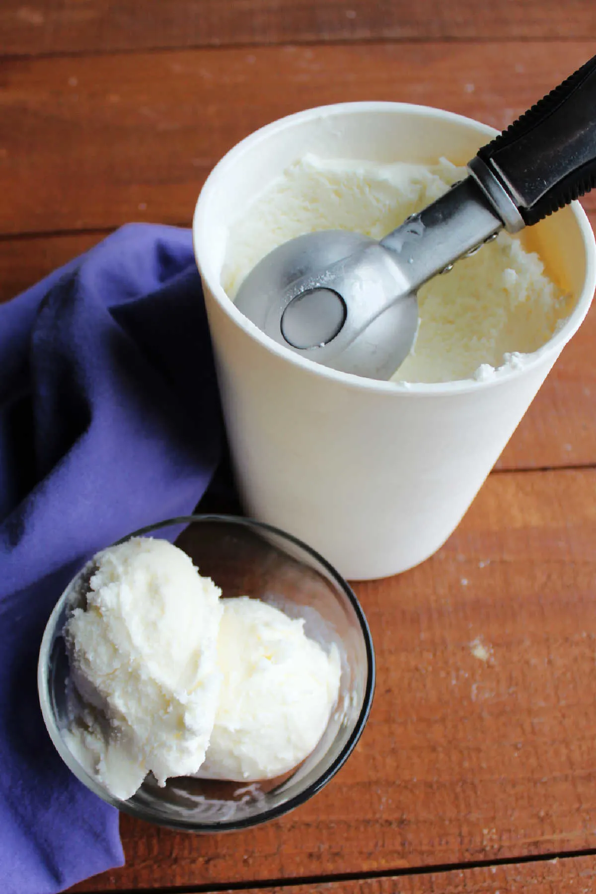 Container of vanilla ice cream with ice cream scoop next to bowl of ice cream. 