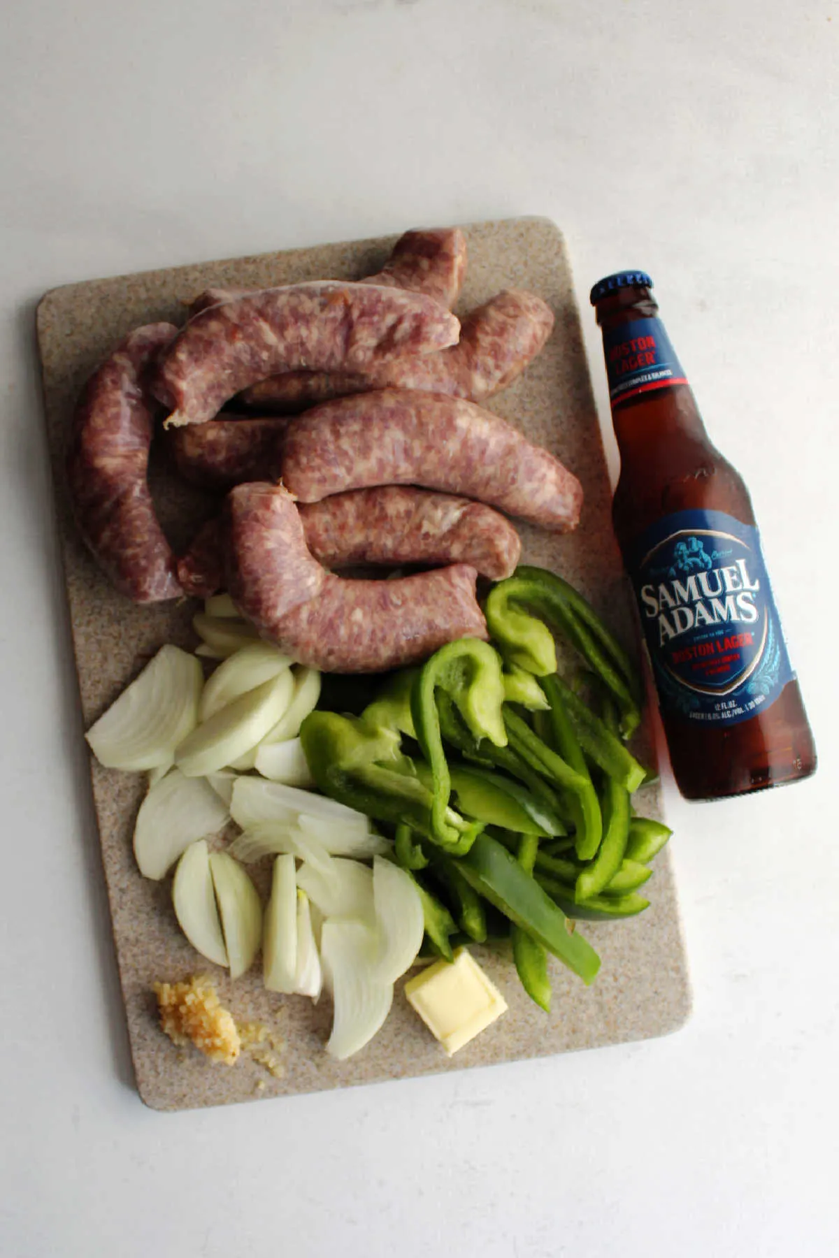 Cutting board filled with sliced onions and peppers, diced garlic, butter and brats next to a bottle of beer.