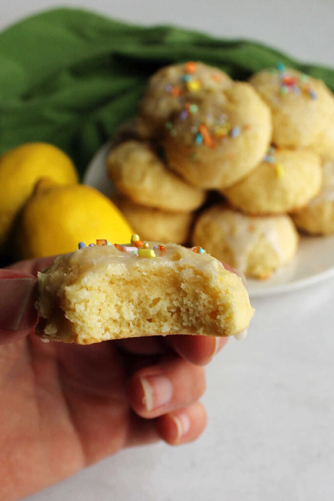 Hand holding iced lemon cookie with a bite taken out showing soft airy interior. 