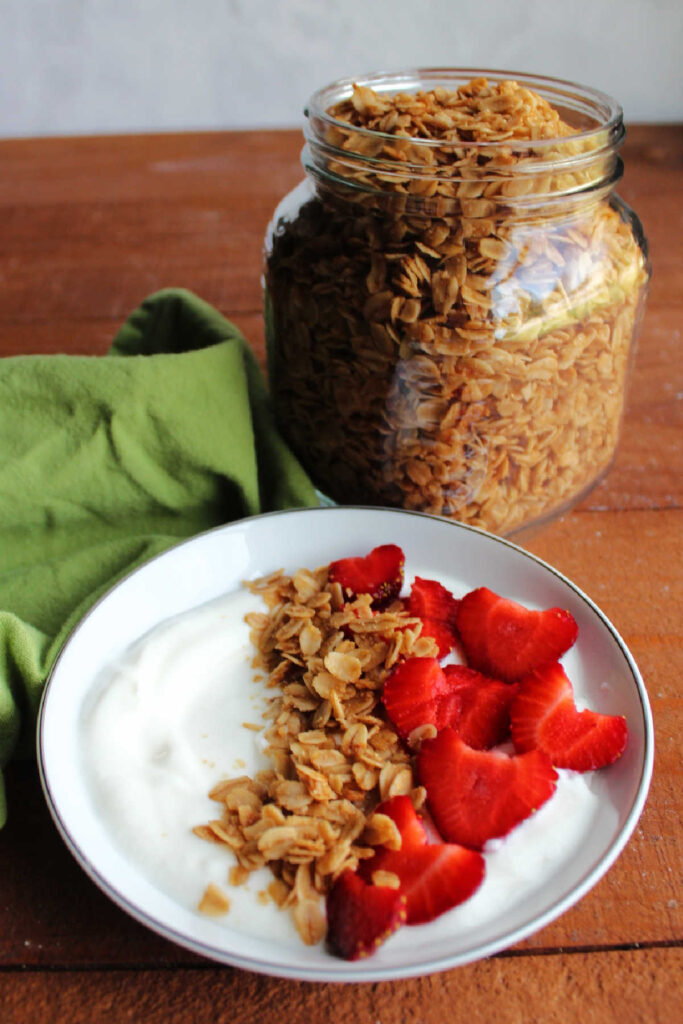 Bowl of yogurt topped with granola and strawberries by jar of maple cinnamon granola.