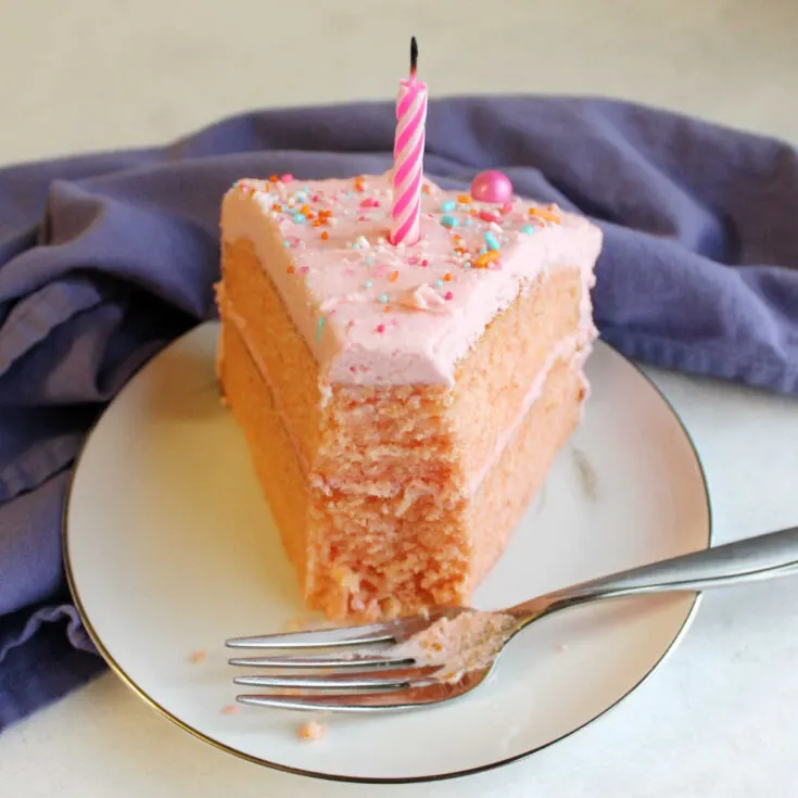 Big slice of pink velvet cake on plate with a bite taken out of the end and a blow out candle on top.