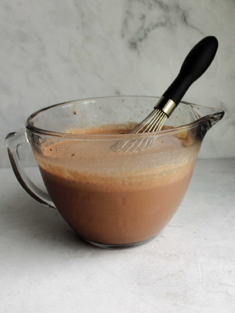 Large glass mixing bowl filled with chocolate ice cream base and whisk.