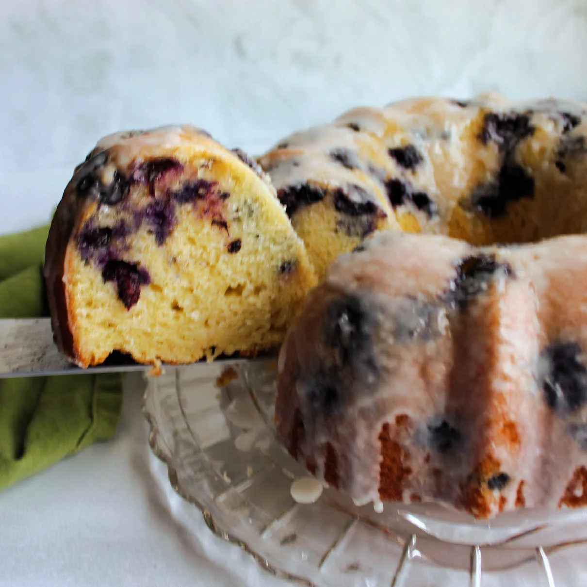 Lifting slice of lemon blueberry bundt cake out of cake showing pale yellow center with lots of blueberry specks.