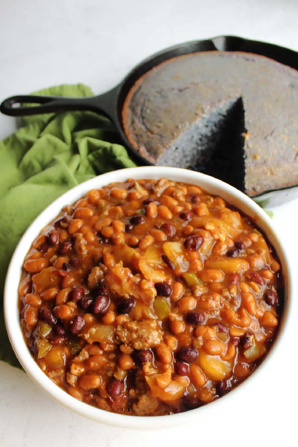 Bowl of loaded baked beans with cast iron skillet of blue cornbread in the background. 