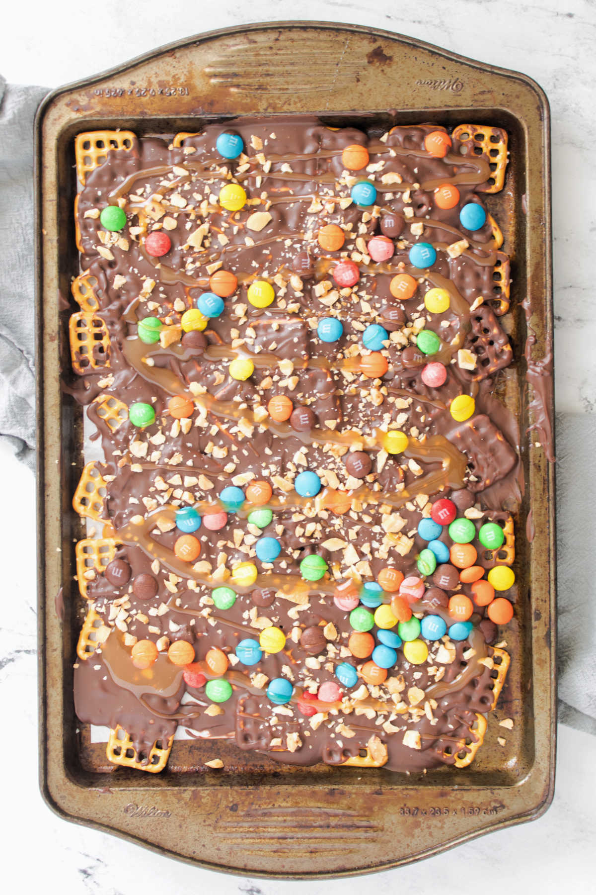 Pan of chocolate bark with pretzels, caramel, chopped peanuts and colorful m&m's. 