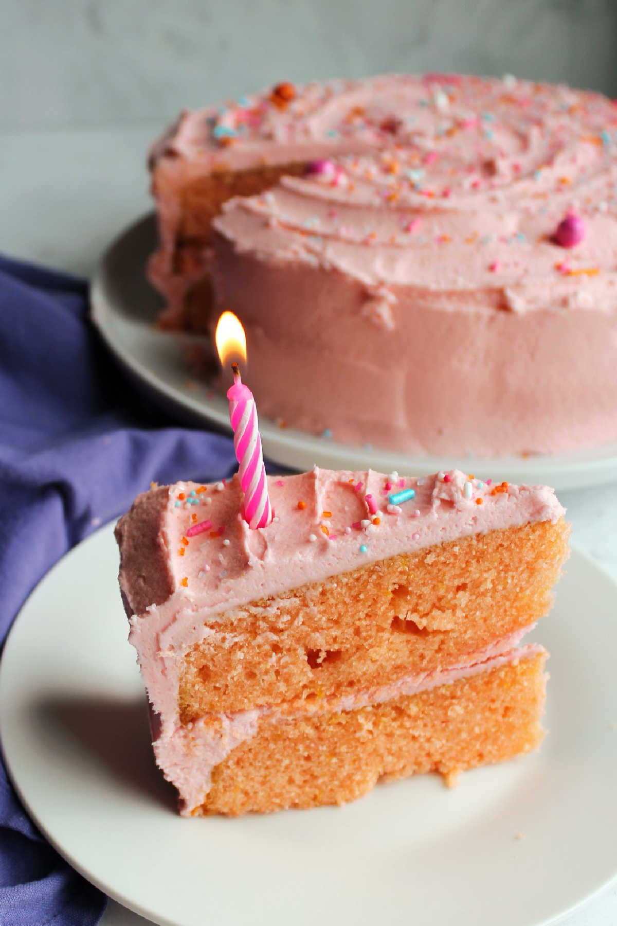 Slice of layered pink velvet cake on plate with lit birthday candle in it. 
