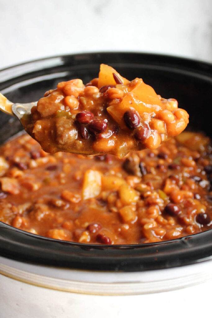 Serving spoon filled with loaded baked beans being lifted out of slow cooker.