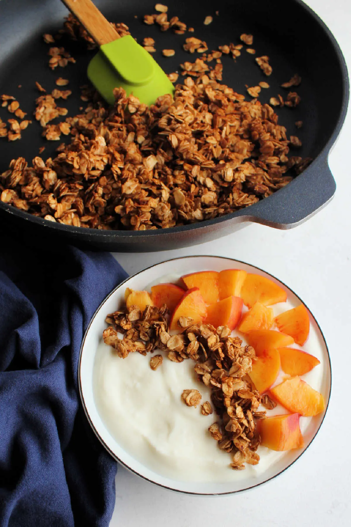 Bowl of yogurt with granola and peaches in front of skillet of remaining granola.