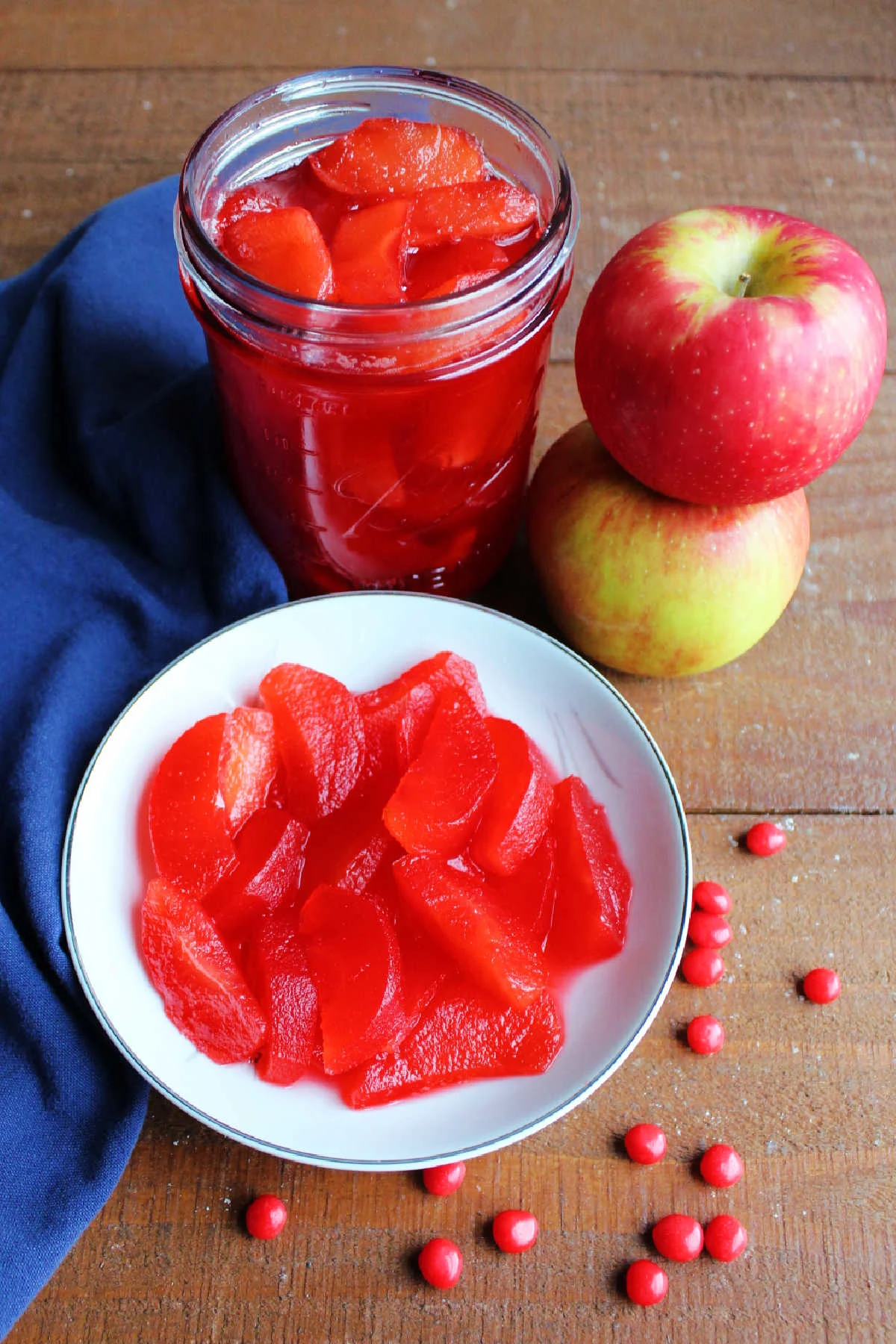 Bowl of red hot apples next to a jar with more apple slices in it with fresh apples and red hot candies.
