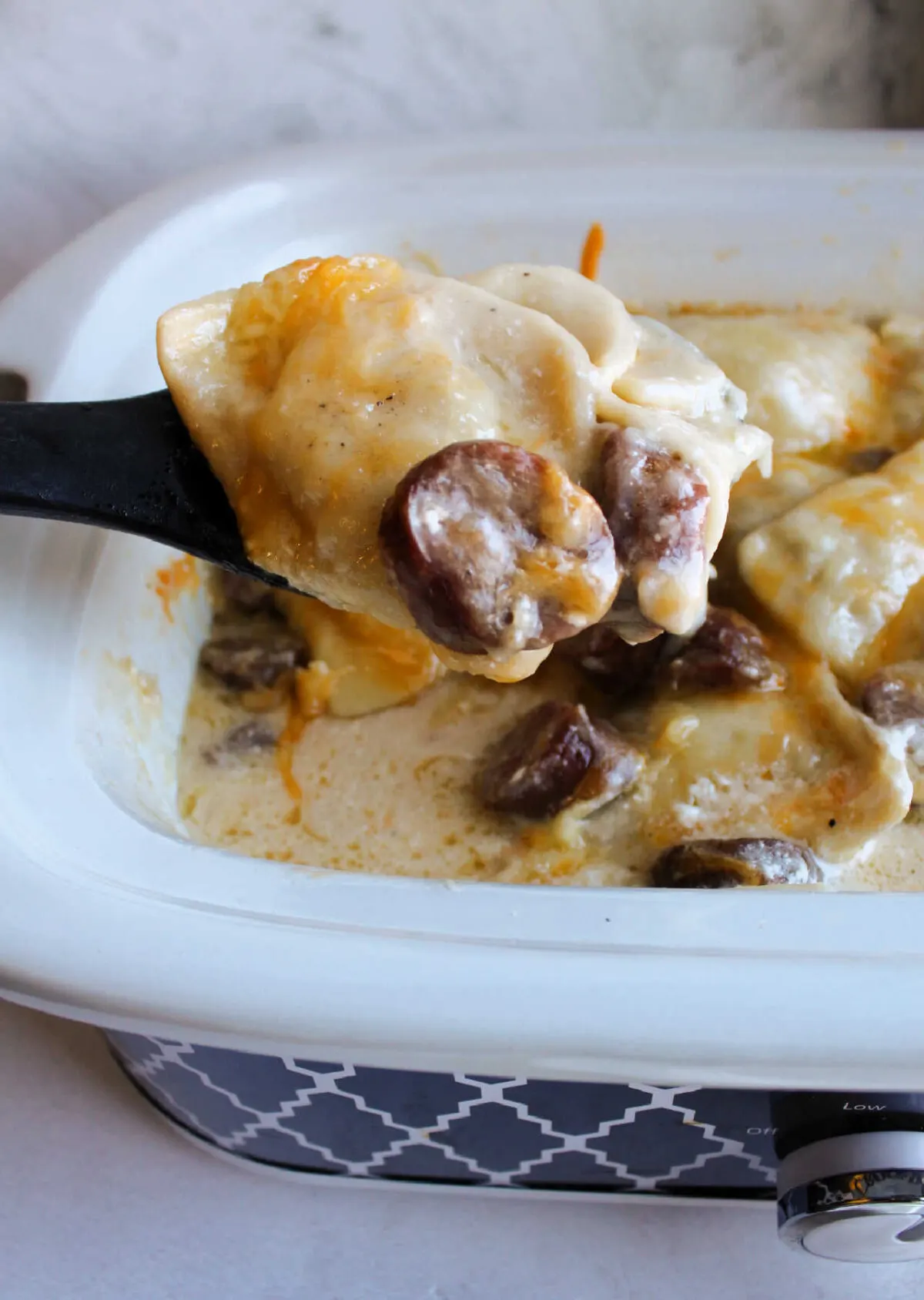 Spoon lifting helping of cheesy pierogies and sausage out of slow cooker.