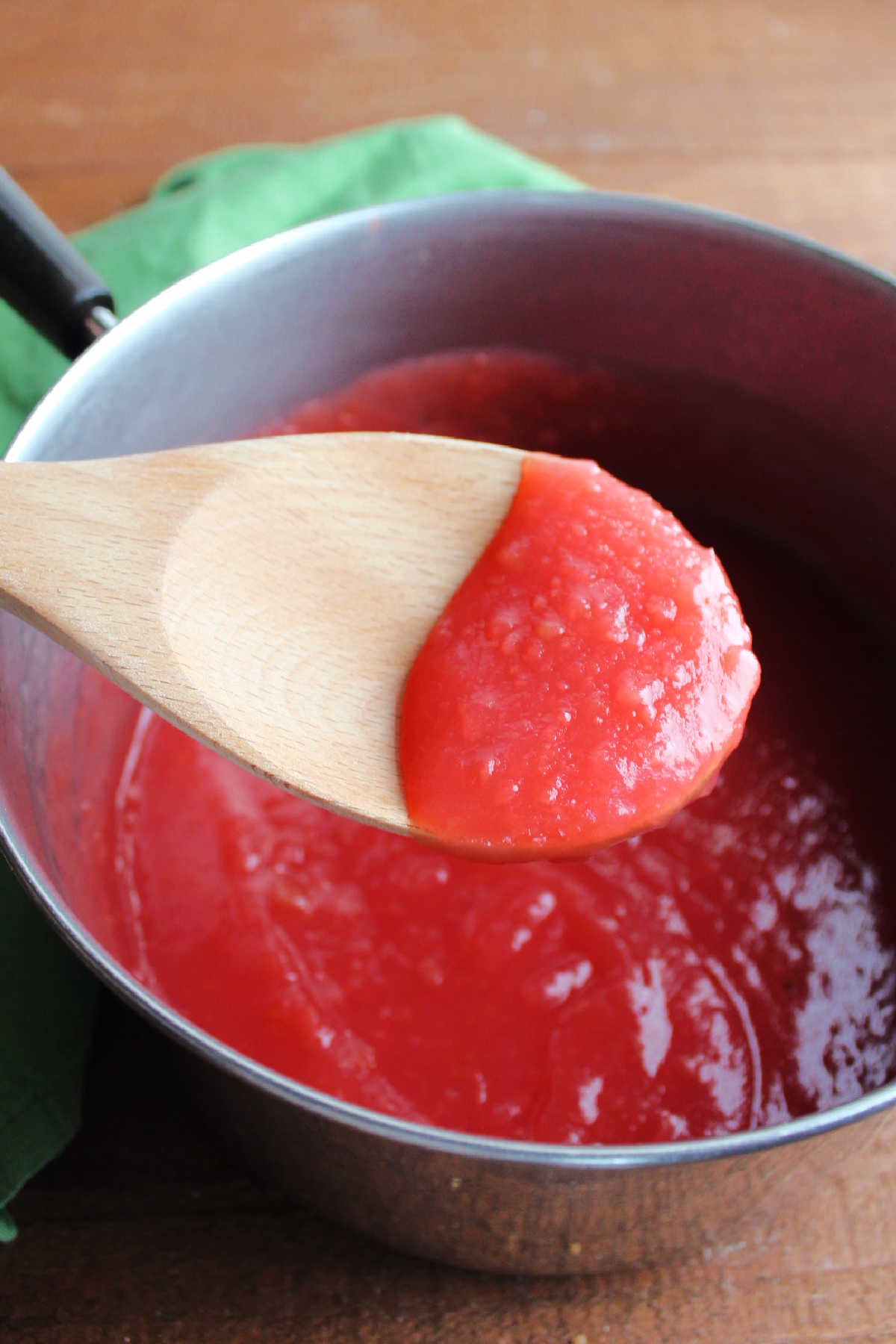 Wooden spoon with freshly made red hot applesauce over saucepan of sauce. 