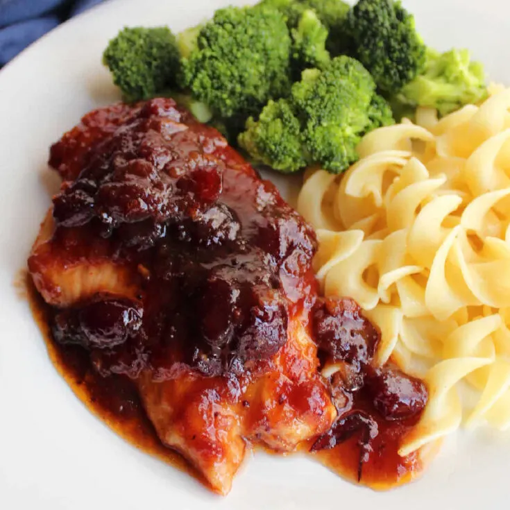 Close up of chicken breast topped with sauce made with whole cranberries and catalina dressing served with pasta and broccoli.