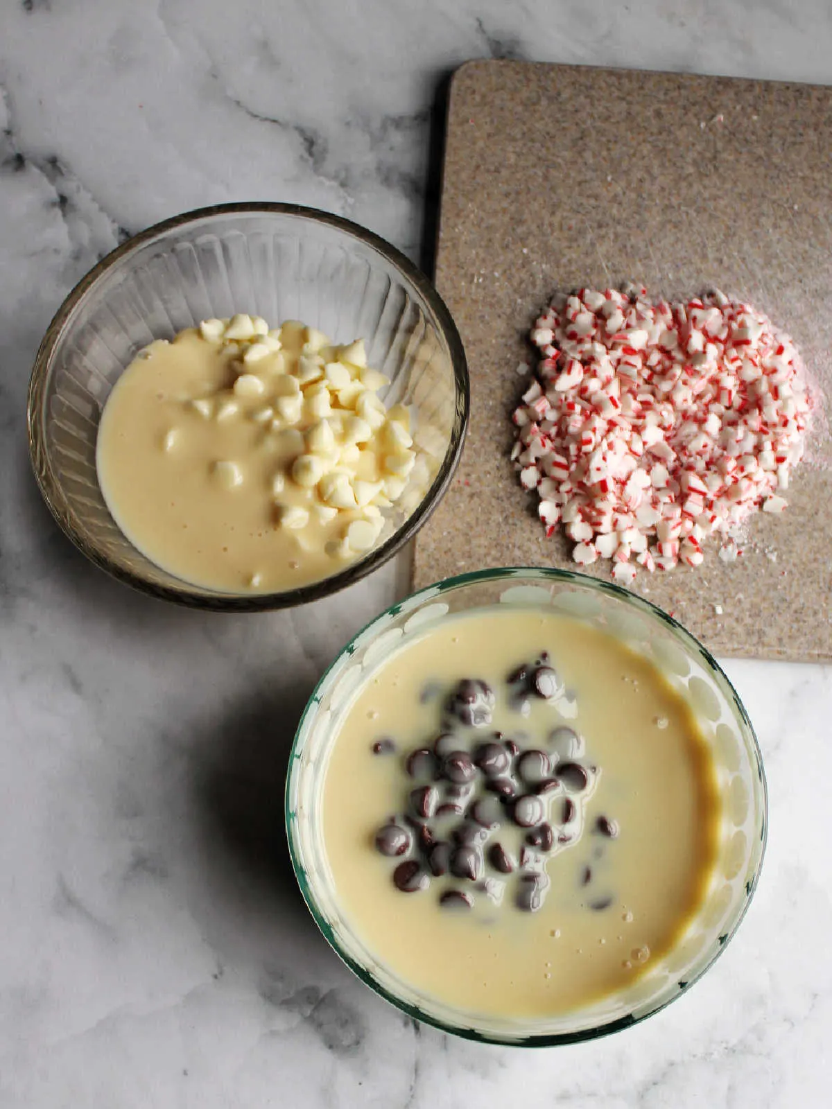 Condensed milk divided between two bowls along with the chocolate chips and white chocolate chips with chopped candy canes on cutting board. 