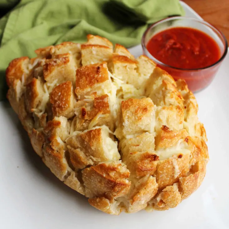 Loaf of cheesy garlic pull apart bread on plate with bowl of marinara sauce for dipping.