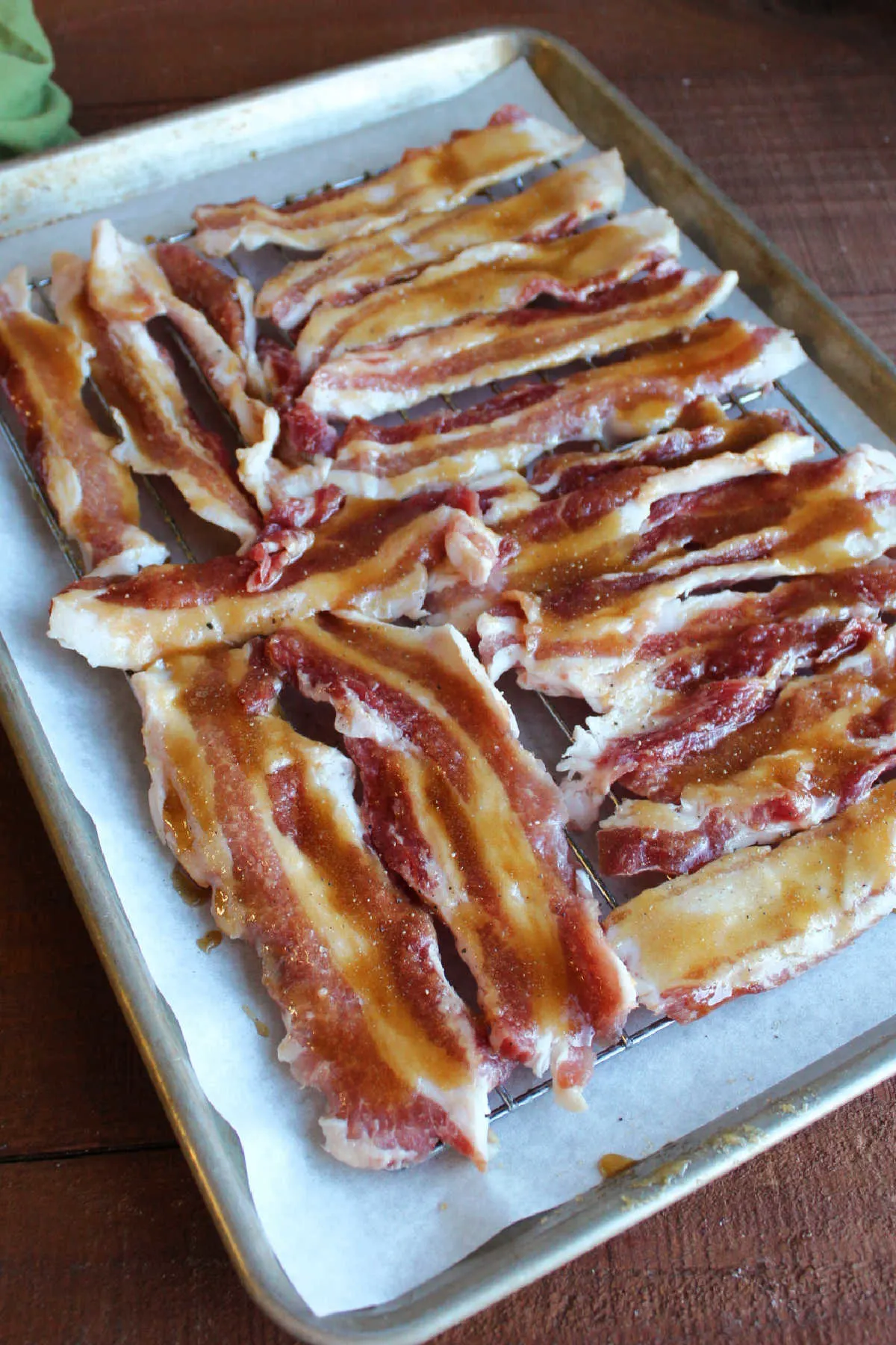 Raw bacon strips coated in maple and brown sugar mixture arranged on wire rack over parchment lined rimmed baking sheet.