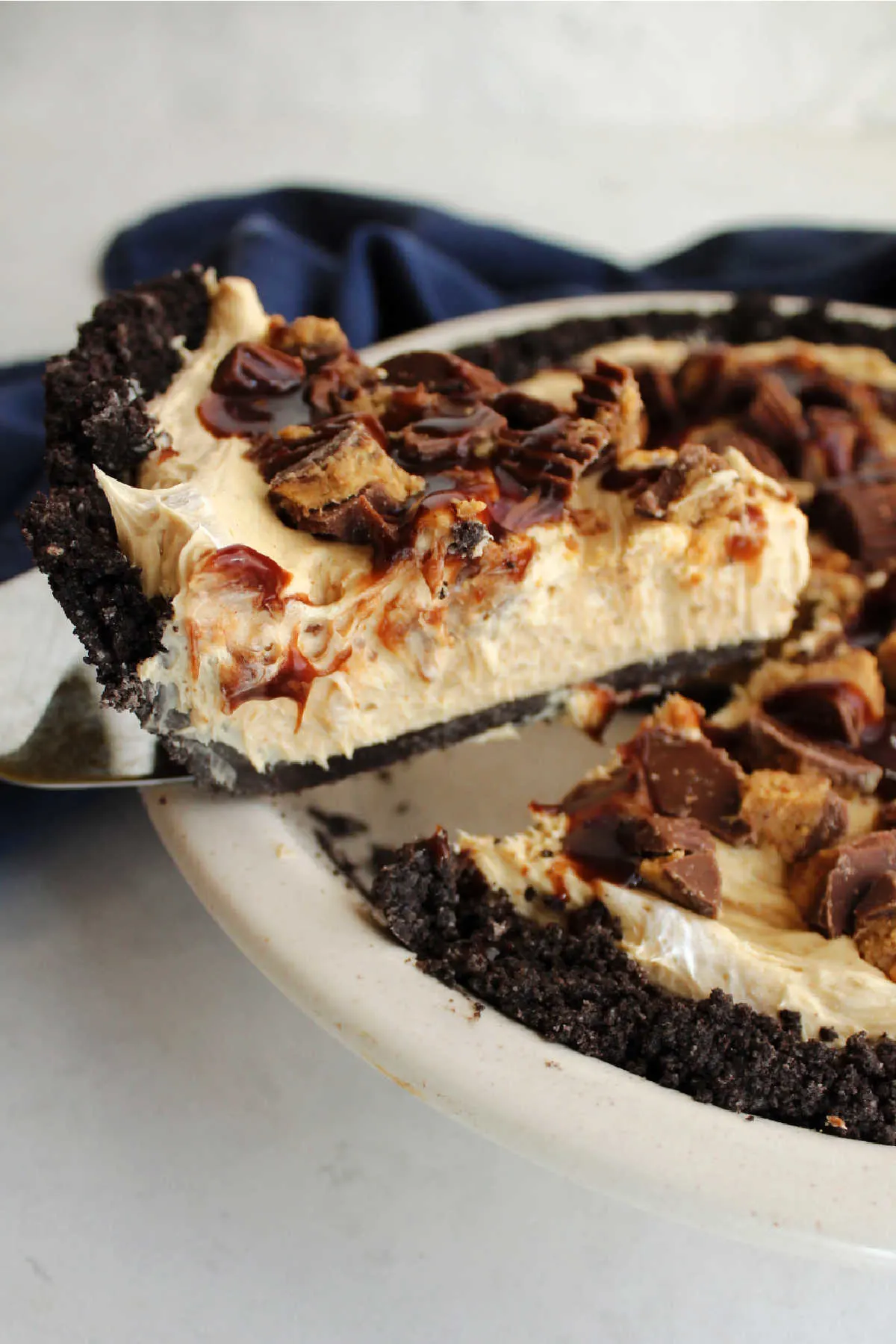 Lifting slice of peanut butter pie out of the pan showing Oreo crust, creamy peanut butter filling and candies and fudge on top.