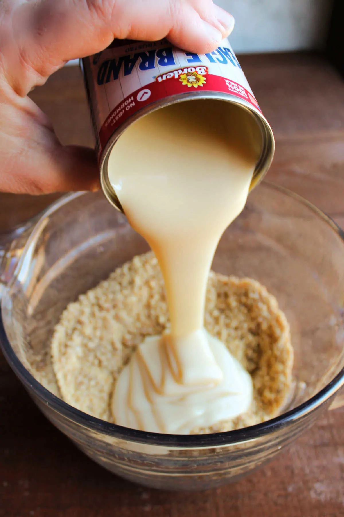 Pouring sweetened condensed milk over graham cracker crumbs in bowl.