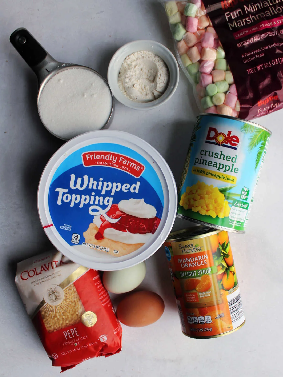 Ingredients including pepe pasta, eggs, crushed pineapple, sugar, flour, pastel marshmallows and a can of mandarin oranges ready to be made into easter frog egg salad.