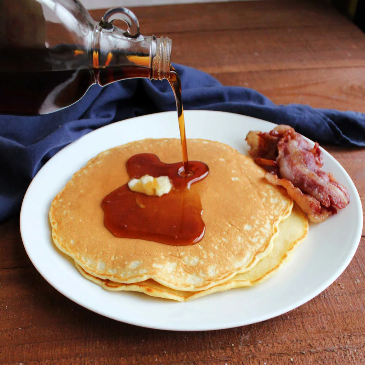 Pouring maple syrup over butter topped homemade pancakes served on plate with strips of bacon.