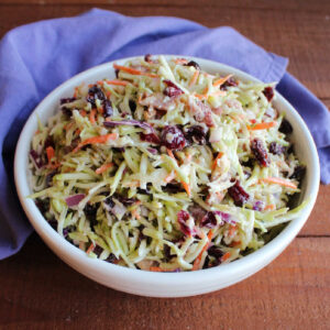 Bowl of broccoli slaw with shaved carrots, dried cranberries, and bacon in a creamy dressing.