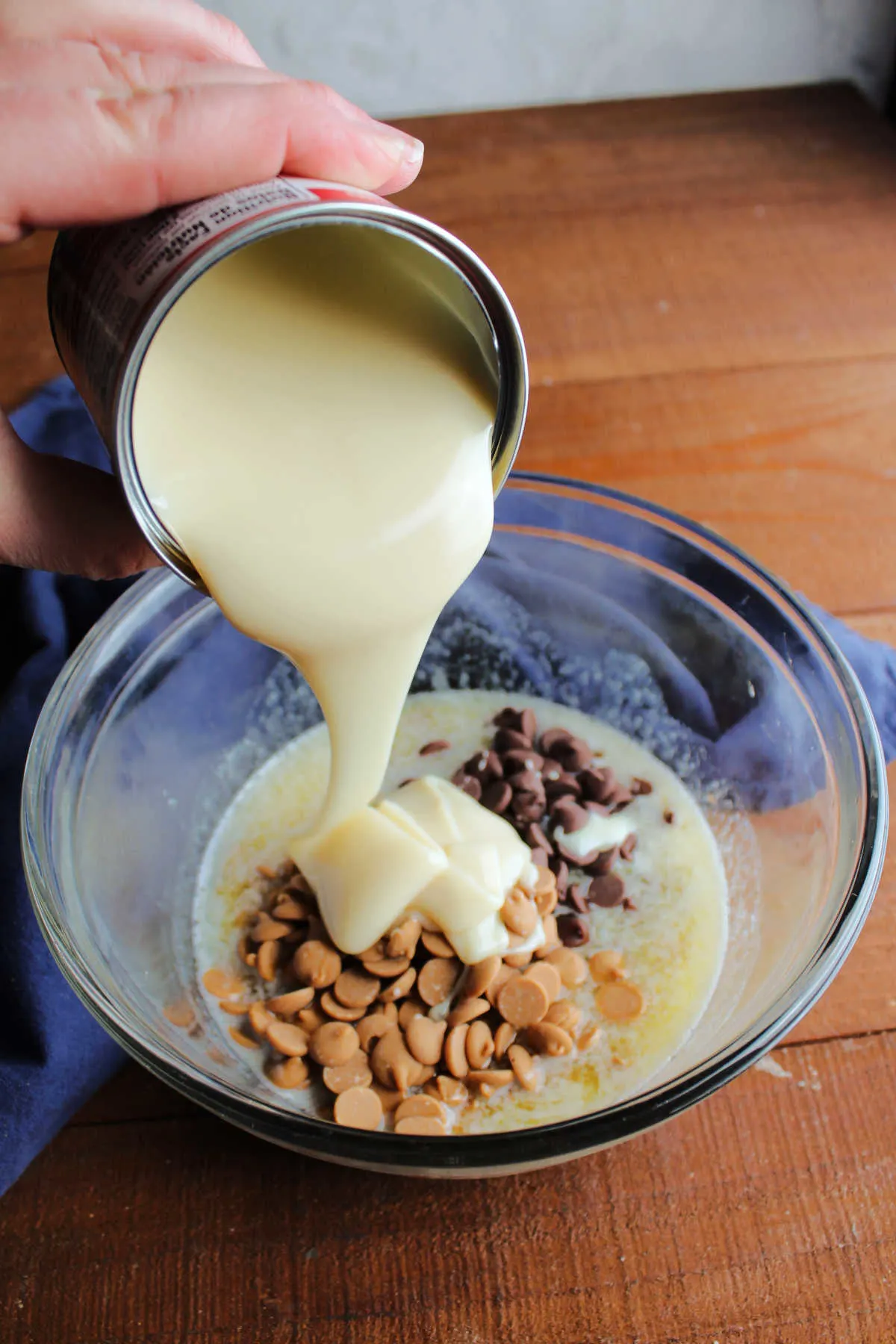 Pouring sweetened condensed milk over bowl of peanut butter chips, chocolate chips, and melted butter.