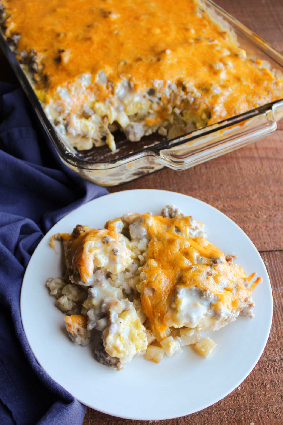 Serving of breakfast casserole with hash browns, eggs, sausage gravy and cheese on plate with remaining casserole in pan behind it.