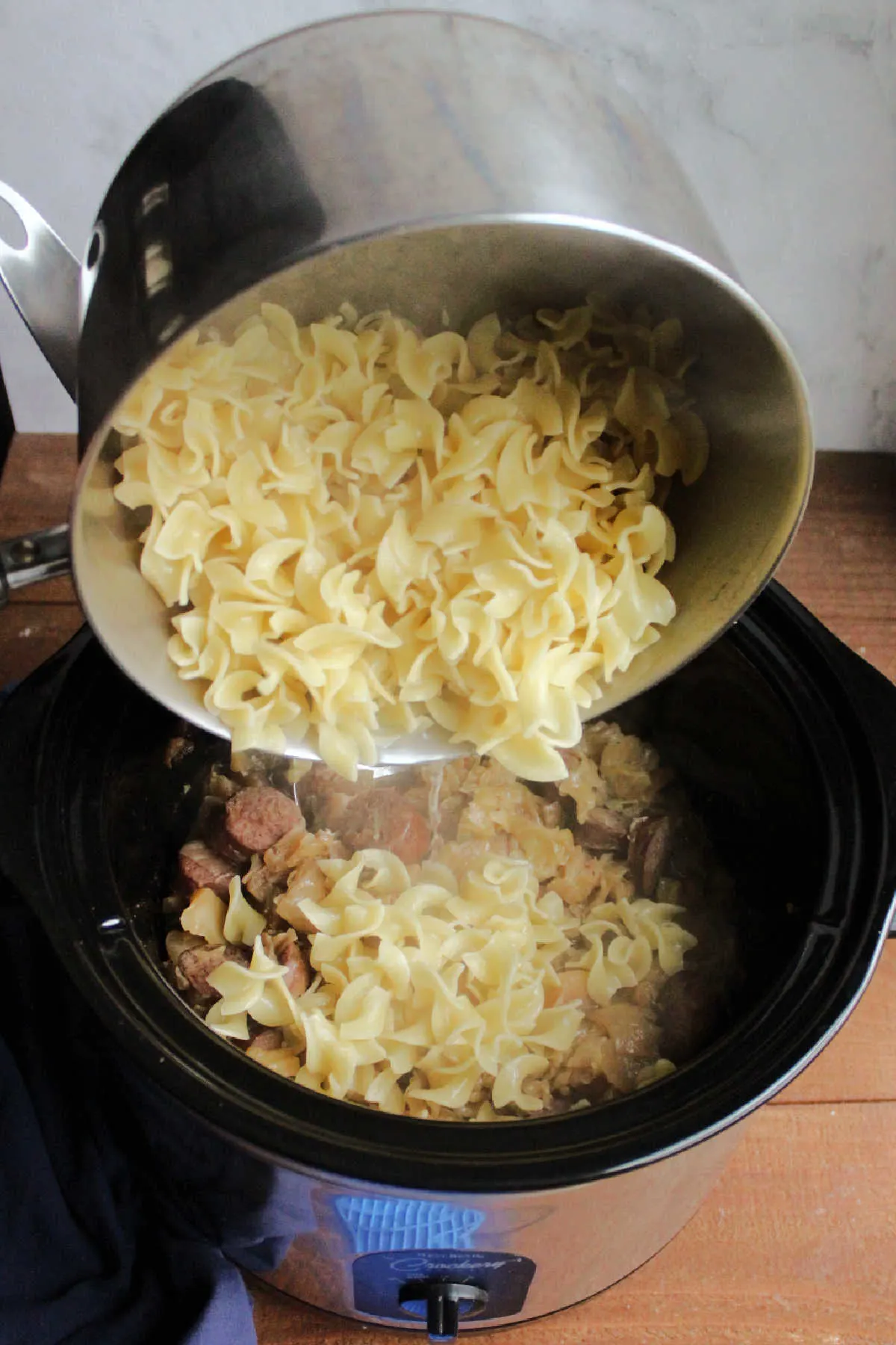 Pouring egg noodles into crockpot with cooked cabbage and kielbasa.