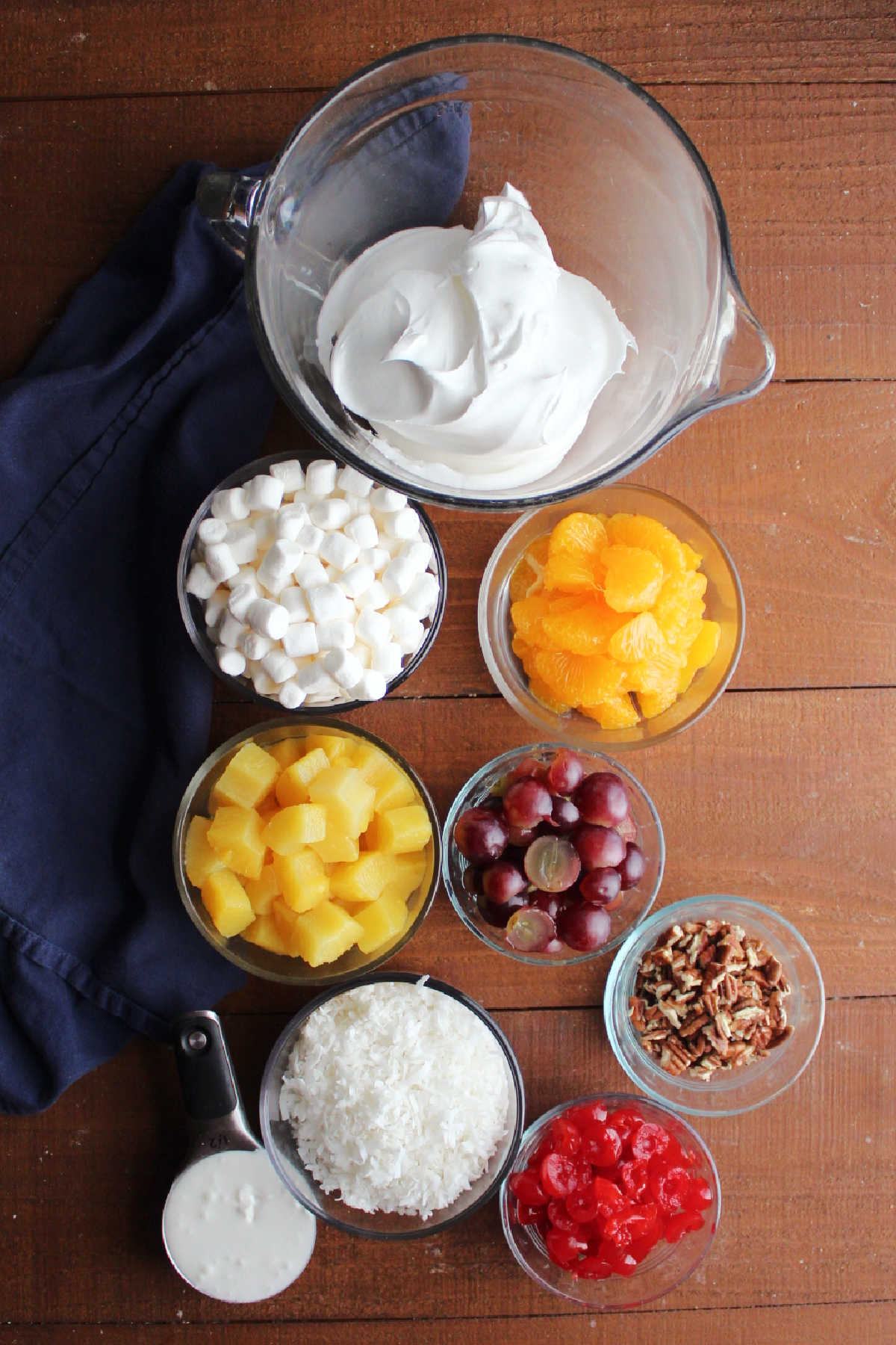 Ingredients including pineapple, oranges, grapes, cherries, pecans, coconut, sour cream, marshmallows, and whipped topping ready to be made into ambrosia. 