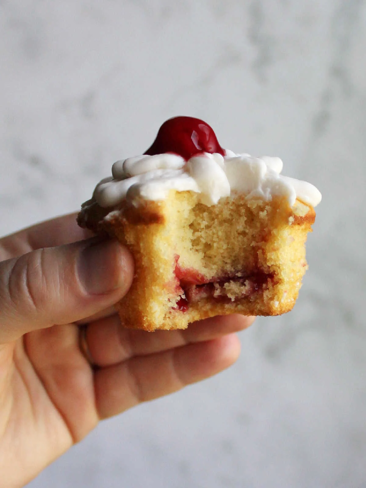 Hand holding cupcake with a bite missing, showing layer of cherry pie filling baked inside and frosting and more cherries on top.