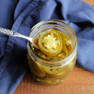 Jar of candied jalapenos with a small pickle fork lifting a pepper ring out of the jar.