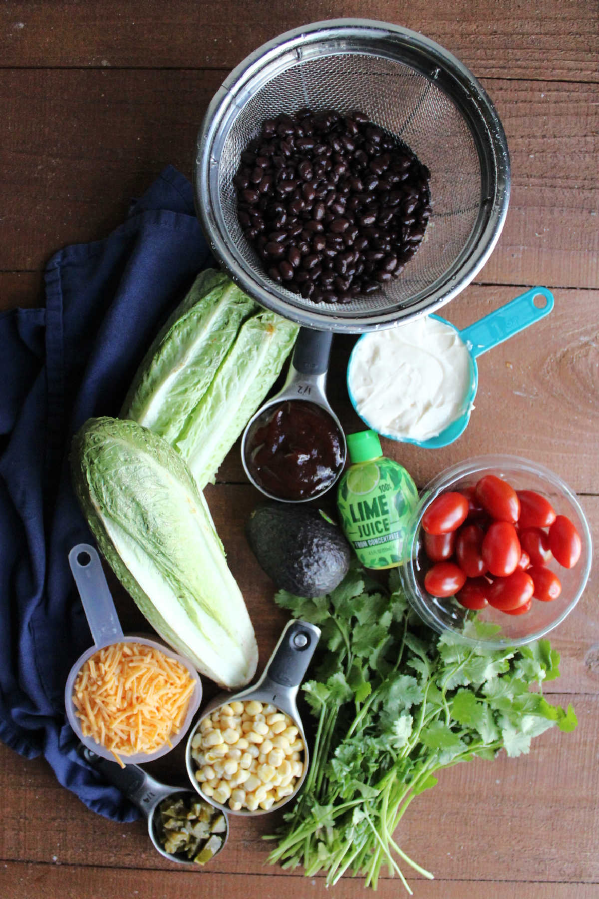 Ingredients including romaine lettuce, black beans, cherry tomatoes, cilantro, corn, cheese, jalapenos, mayonnaise, bbq sauce, and lime juice ready to be made into cowboy salad.