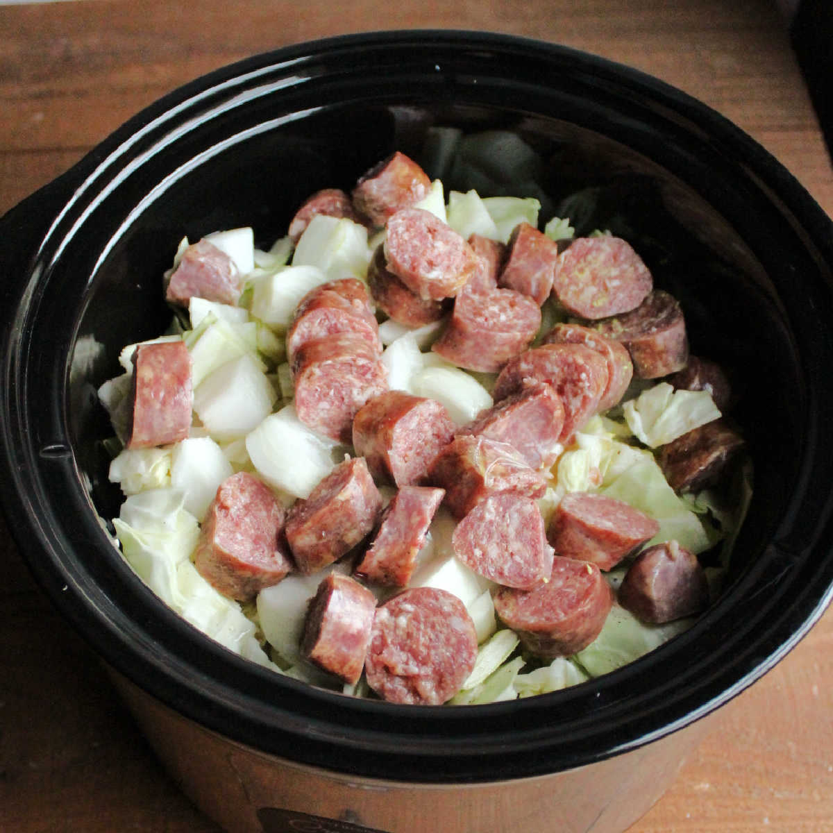 Slow cooker filled with cabbage, onion, kielbasa, seasonings and chicken broth, ready to cook.