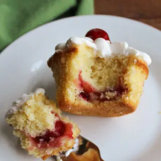 Cupcake on plate with a bite on a fork showing cherry filling inside the cupcake and white frosting and more cherries on top.