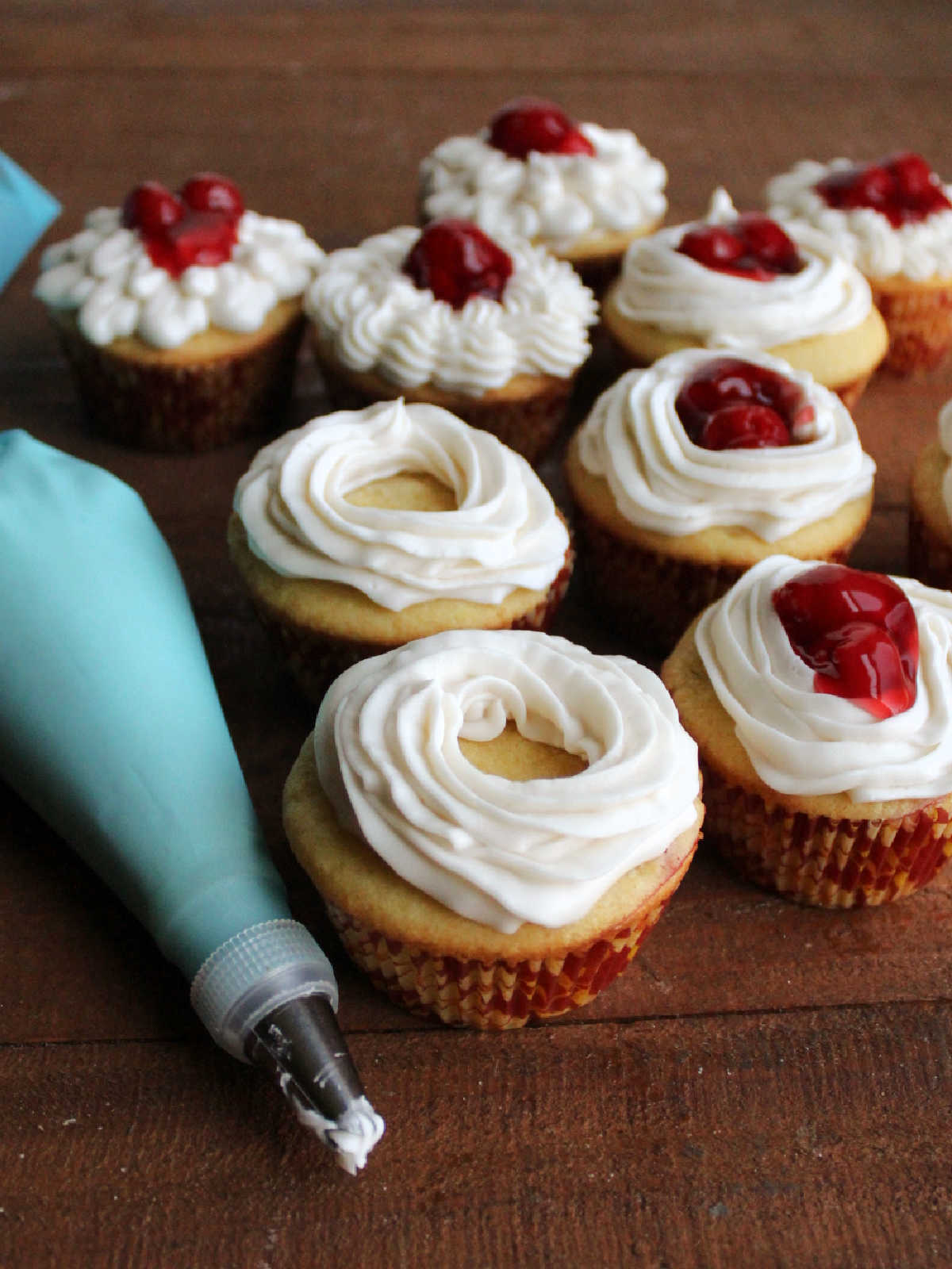 Piping bag with vanilla buttercream next to cupcakes with buttercream rings piped on top, some filled with cherry pie filling.