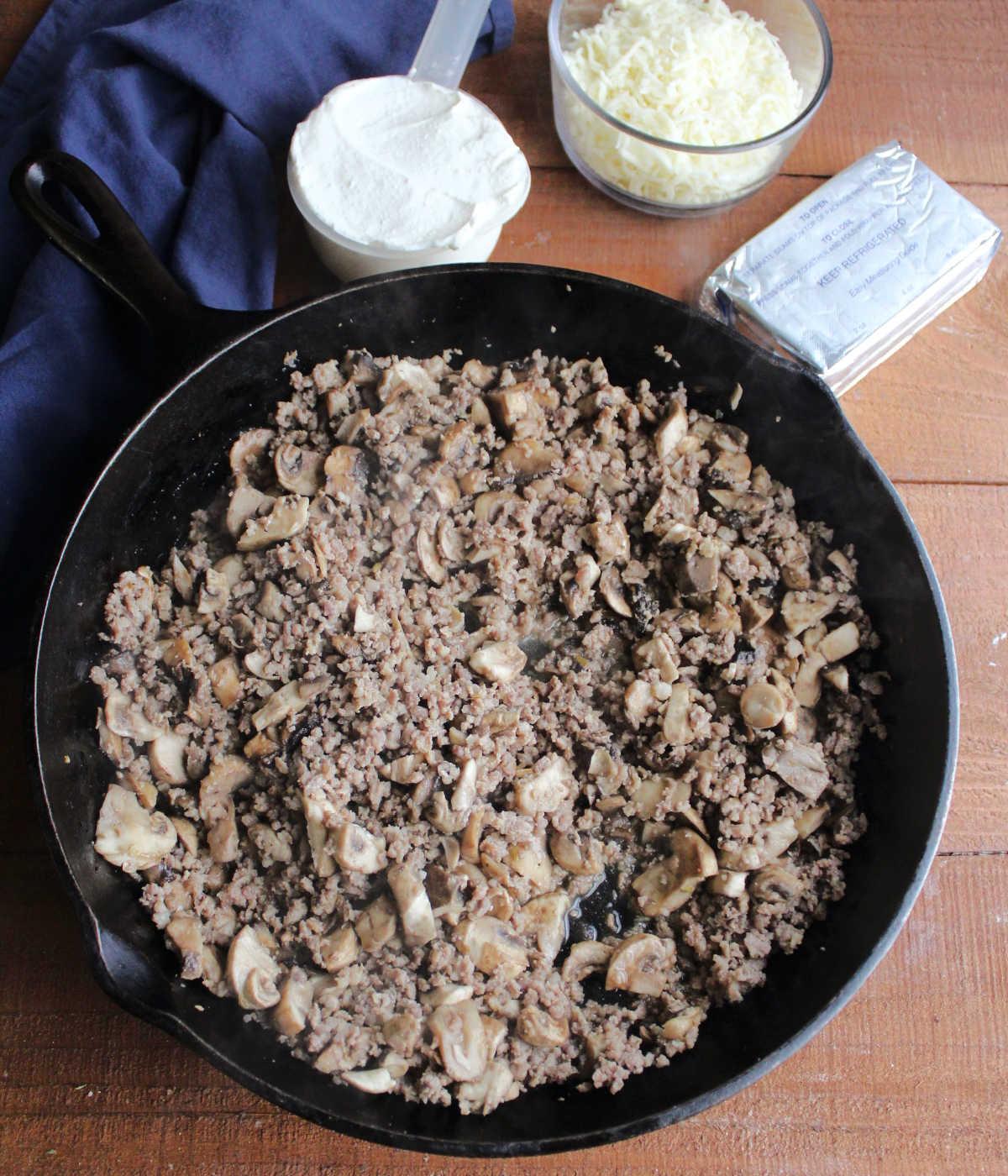 Large cast iron skillet filled with browned sausage and mushrooms next to cream cheese, sour cream, and shredded cheese.
