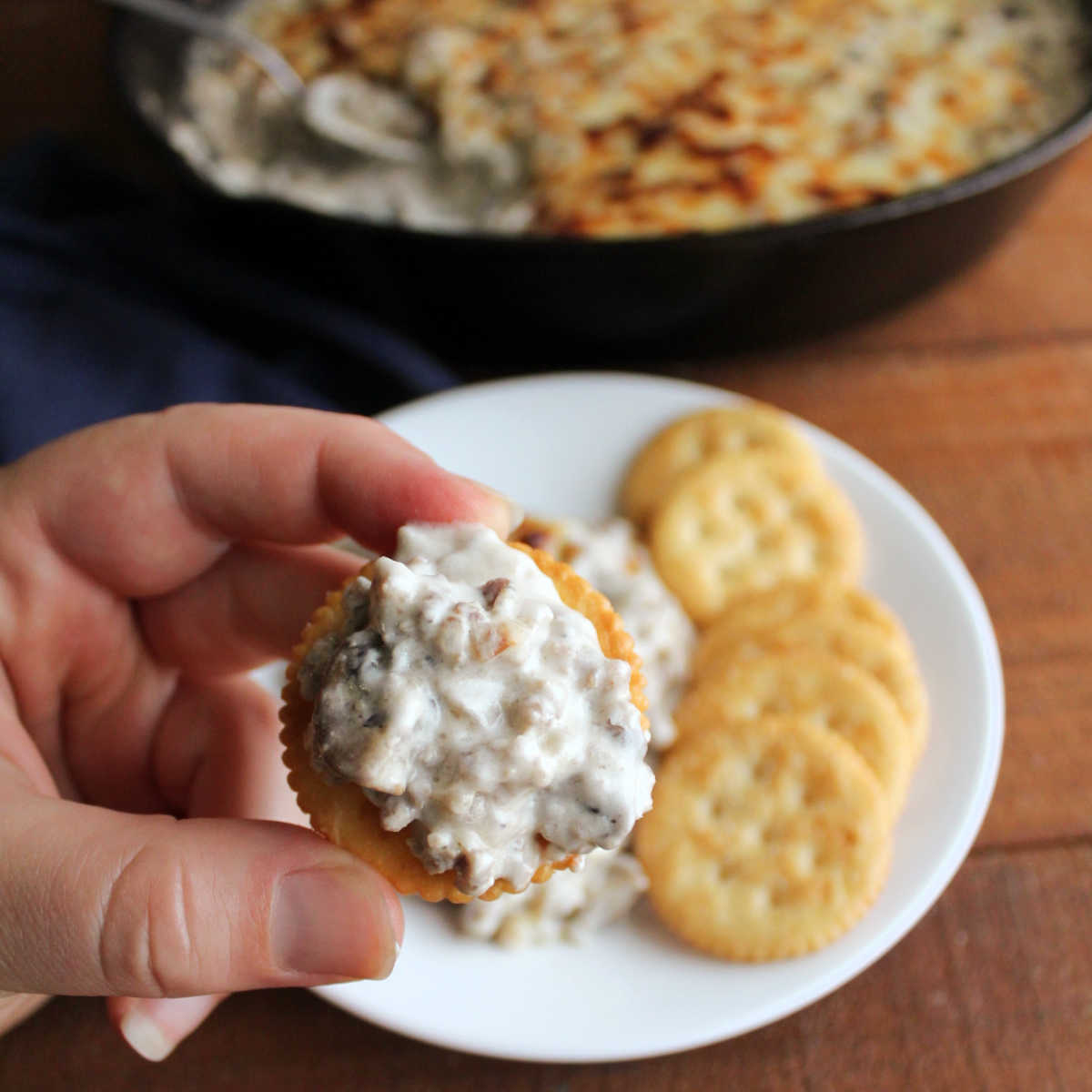 Hand holding cracker topped with creamy stuffed mushroom dip with sausage and cheese.