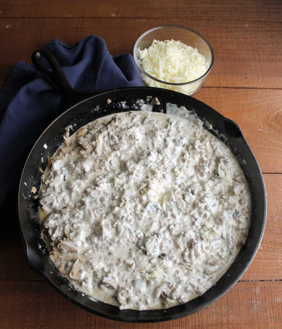 Cast iron skillet filled with creamy stuffed mushroom dip mixture with bowl of shredded cheese nearby.