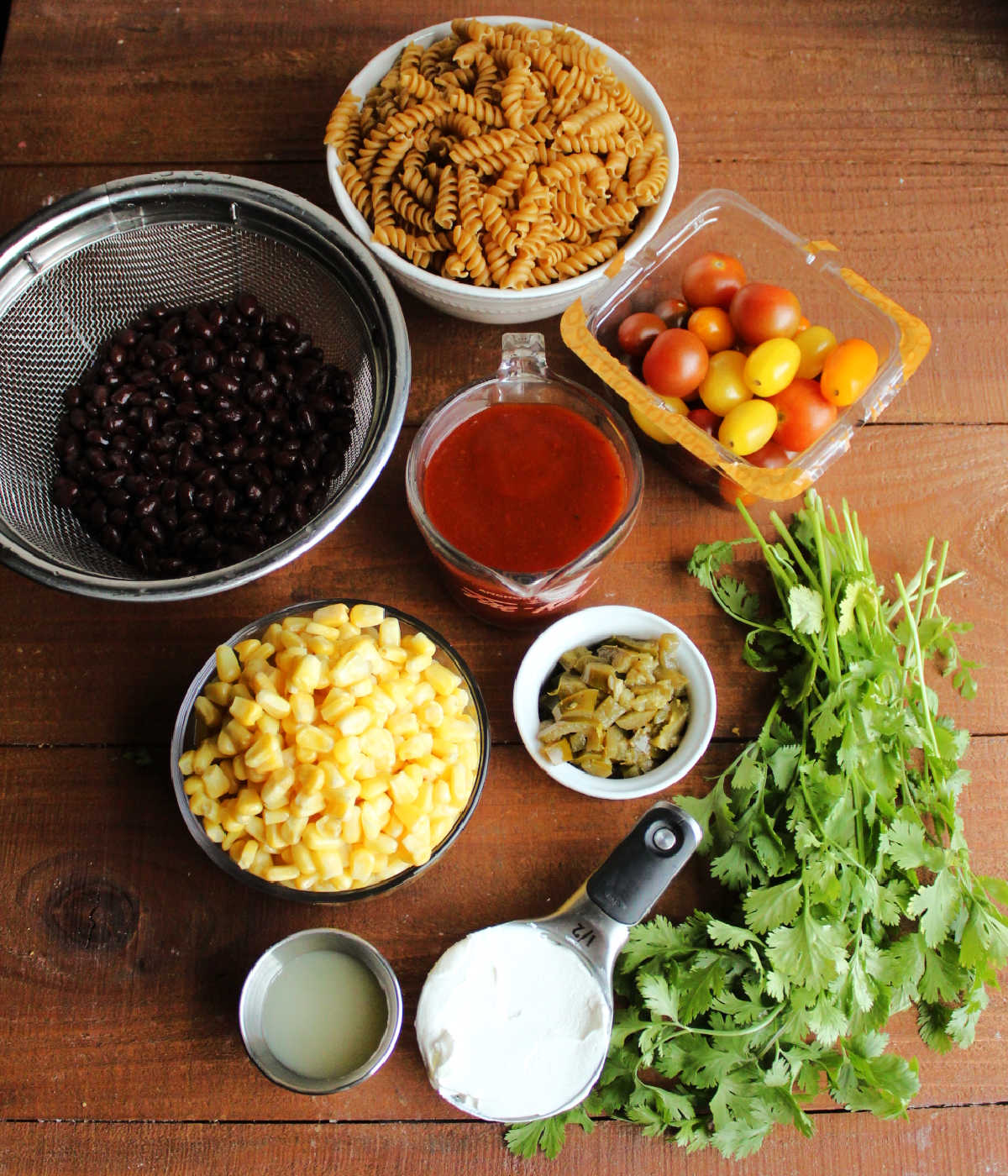 Ingredients including rotini pasta, cherry tomatoes, cilantro. sour cream, lime juice, chopped pickled jalapenos, red enchilada sauce, corn, and black beans ready to be made into pasta salad.