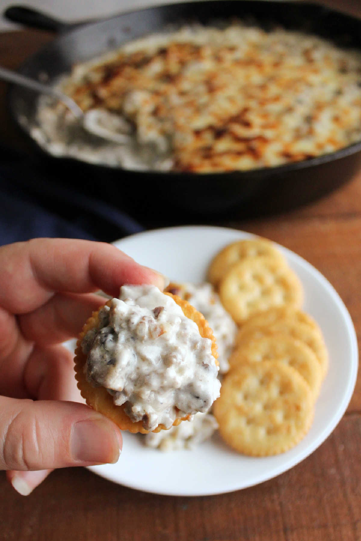 Hand holding cracker topped with cheesy dip with mushrooms and sausage.