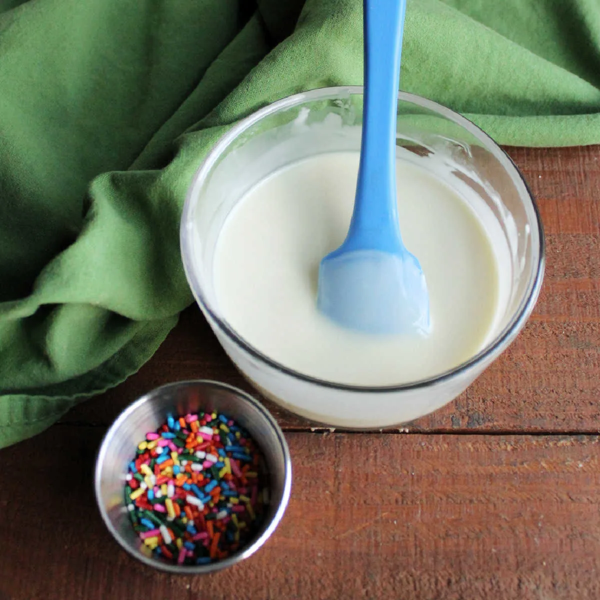 Melted white chocolate and coconut oil in small glass bowl next to a container of colorful sprinkles. 