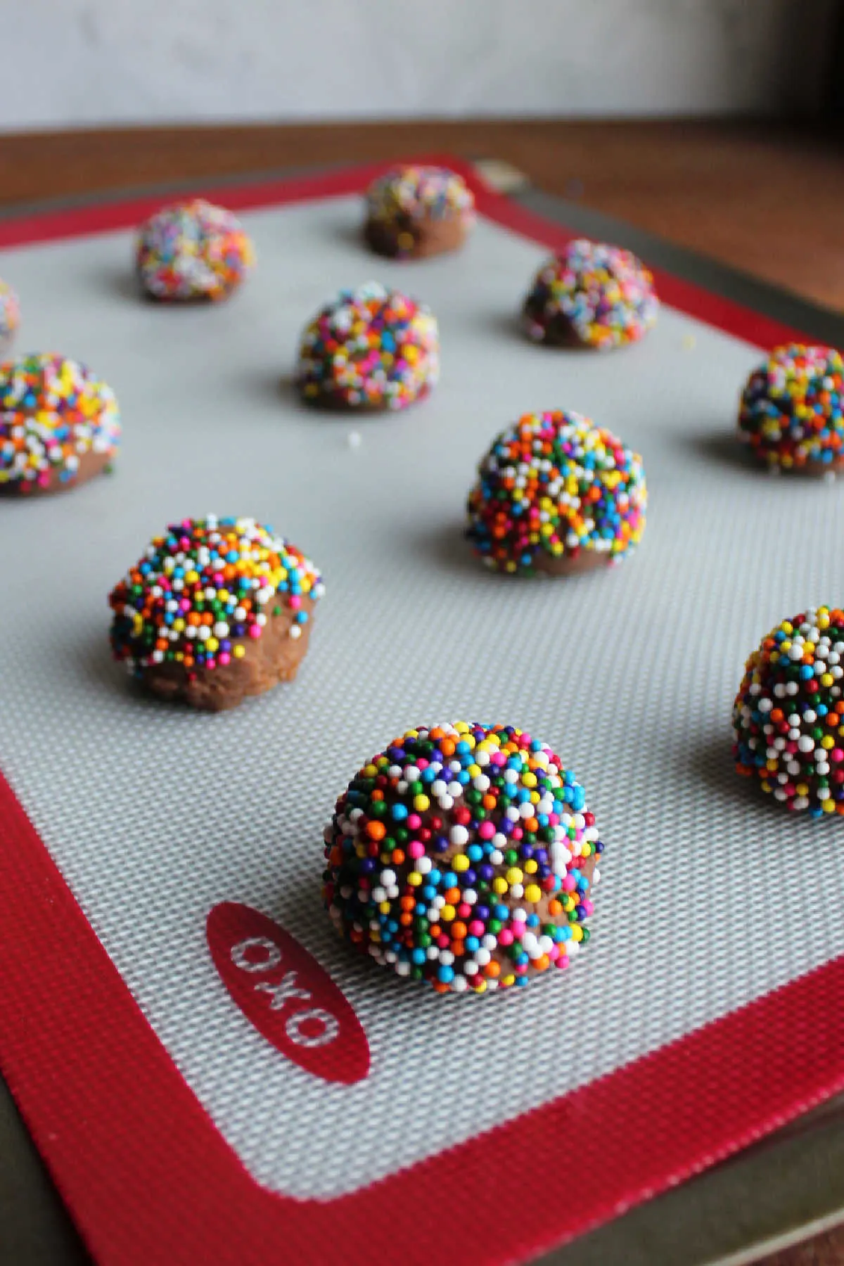 Scoops of chocolate cookie dough rolled in colorful sprinkles on a silicone baking mat covered pan.
