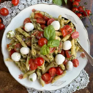 Plate piled high with pesto pasta salad topped with fresh cherry tomatoes, mozzarella pears, and basil.