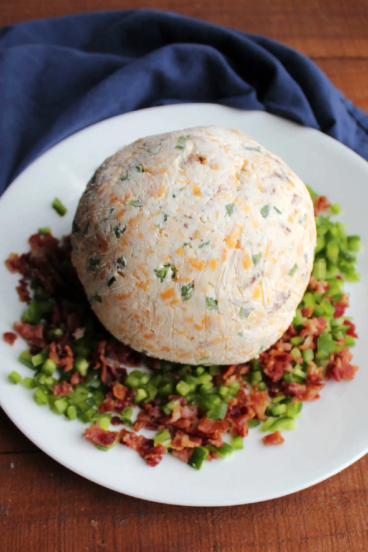 Chilled cheese ball on plate with diced jalapenos and crumbled bacon, ready to be coated and served.
