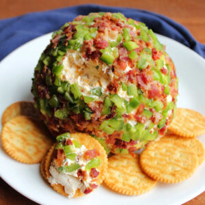 Cheese ball coated in bits of jalapeno pepper and bacon with a little bit missing showing cheesy inside on plate with crackers, ready to eat.