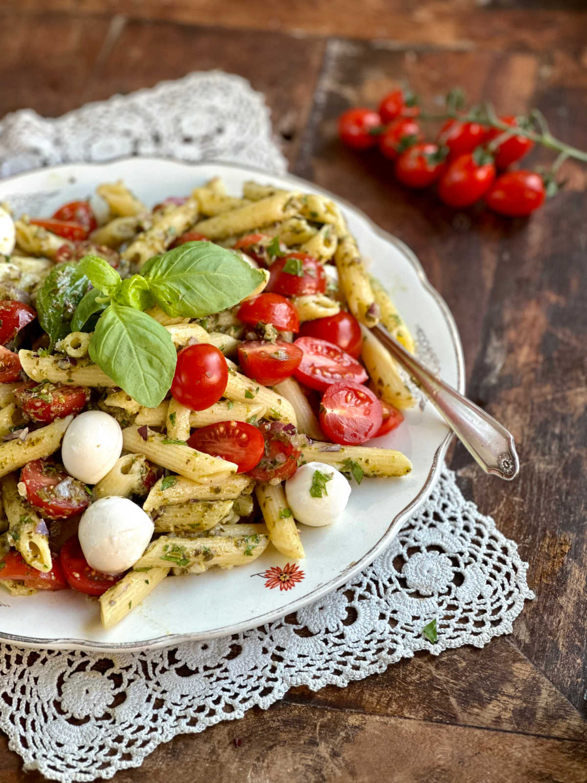 Looking across a plate of pesto penne salad topped with halved cherry tomatoes, mozzarella pearls, and fresh basil, ready to eat.