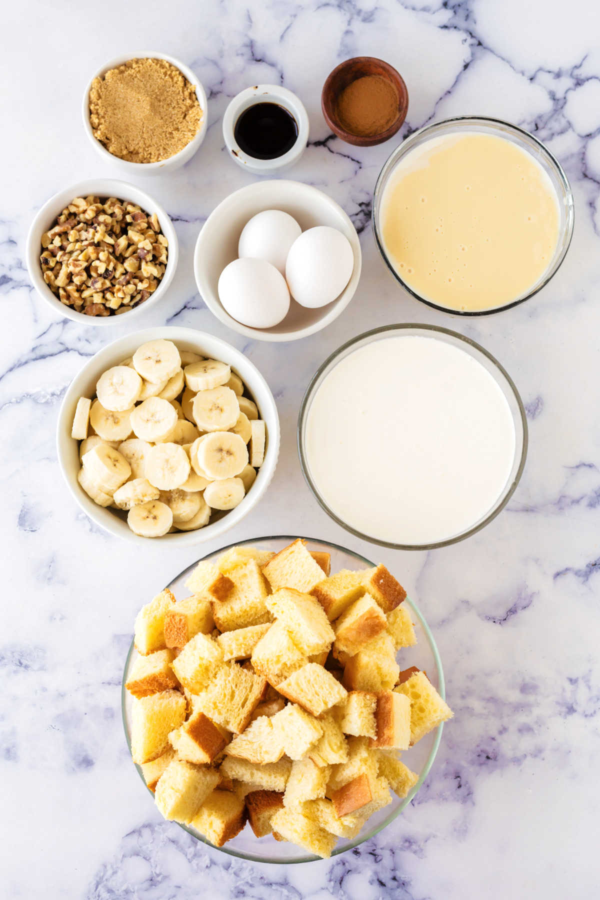 Ingredients including cubed bread, milk, eggs, sweetened condensed milk, bananas, walnuts, brown sugar, vanilla, and cinnamon ready to be made into banana bread pudding.
