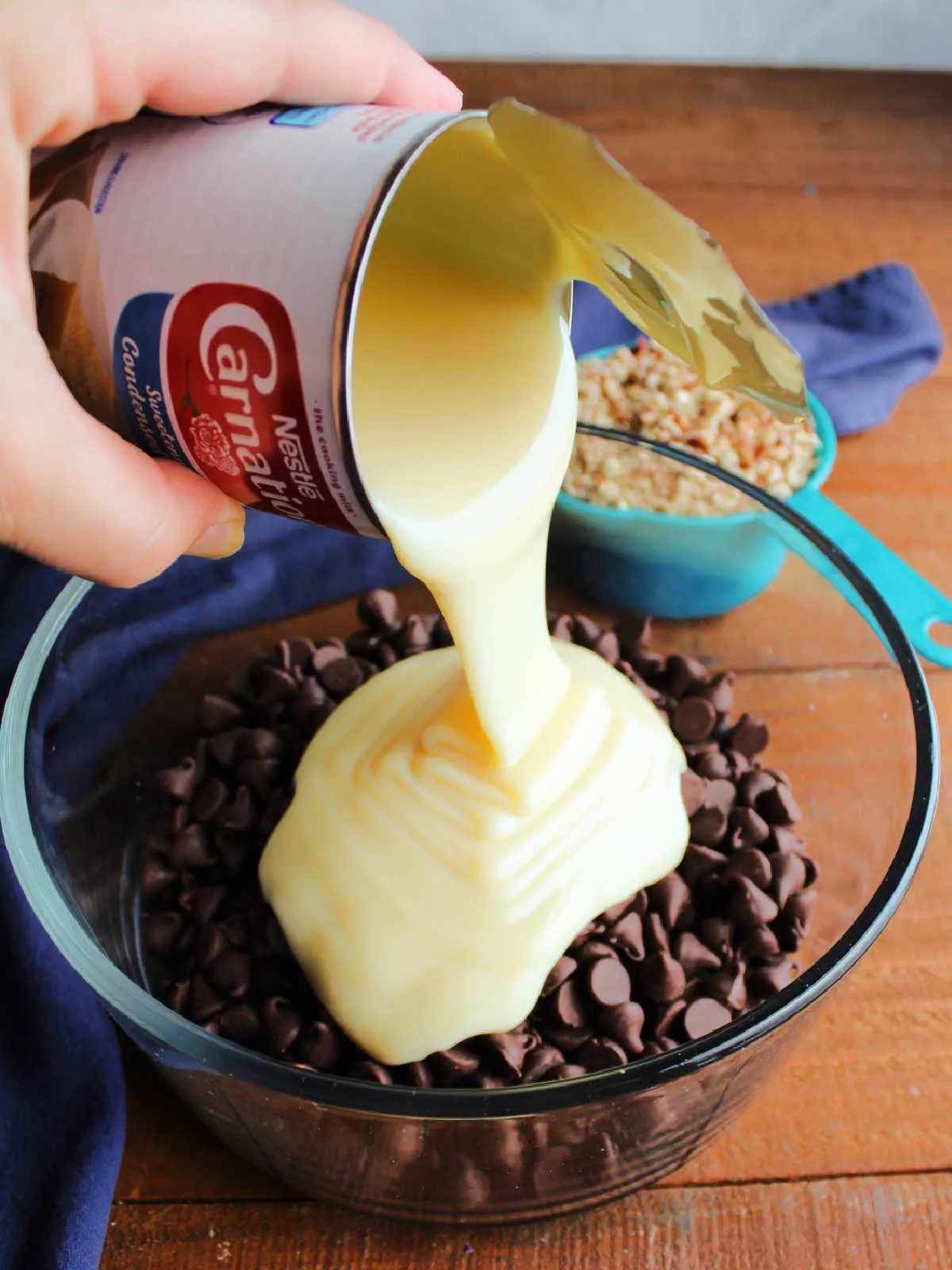 Pouring can of condensed milk over chocolate chips in a glass bowl.