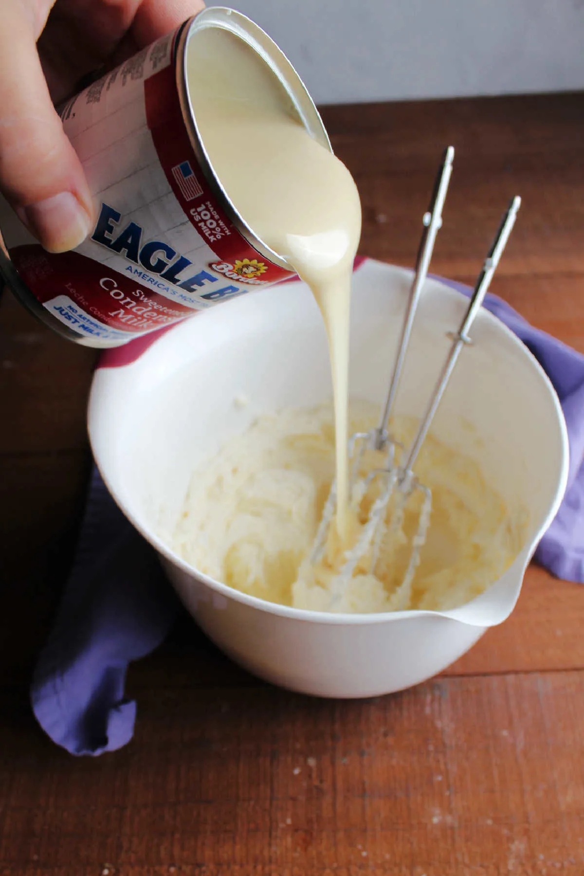 Pouring can of sweetened condensed milk into a mixing bowl with fluffy cream cheese and mixer beaters.