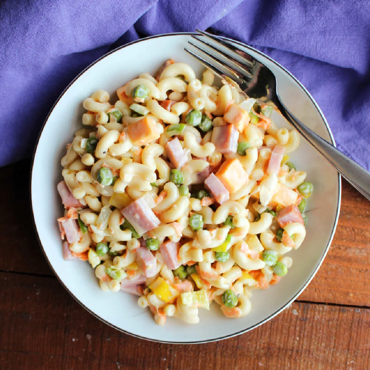 Lunch size portion of sweet pasta salad with peas, ham, cheese, onions, carrots, and peppers in a creamy tangy and sweet dressing with a fork, ready to eat.